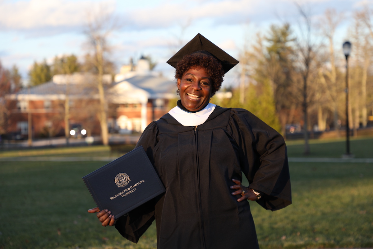 Woman standing outside and smiling in cap and gown holding diploma cover