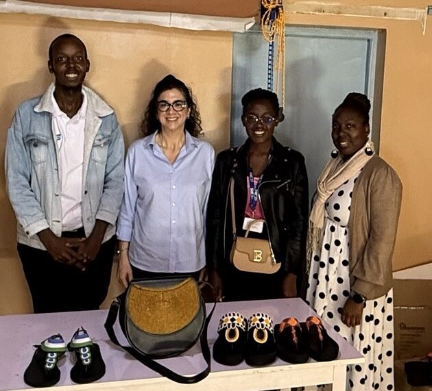 Four people stand behind a table displaying handcrafted items, including several pairs of shoes and handbags, inside a workshop or showroom. The group smiles at the camera while showcasing the products, with shelves and workshop materials visible in the background.