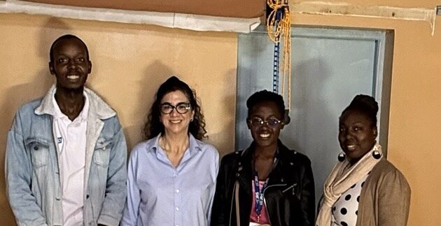 Four people stand behind a table displaying handcrafted items, including several pairs of shoes and handbags, inside a workshop or showroom. The group smiles at the camera while showcasing the products, with shelves and workshop materials visible in the background.