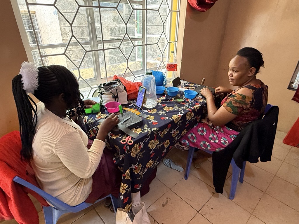 Two women sit at a table by a large window, working on a craft project. The table is covered with tools, materials, and containers, while one woman focuses on assembling an item and the other looks toward the camera. The scene depicts a small-scale workshop or training session in a bright indoor setting.