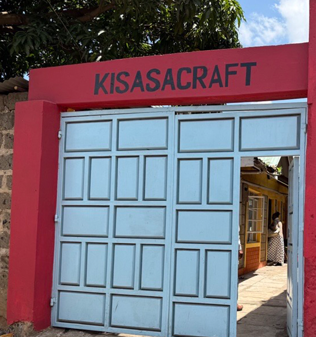 The entrance gate to Kisasacraft, featuring a red sign with the organization’s name above large light-blue metal doors. Through the open gate, part of the workshop compound and a person walking inside can be seen.